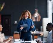 A female standing talking to a group of people around a table